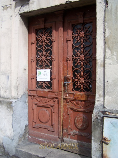 Stare drzwi z kratami ozdobnymi, które posiadają wszystkie połączenia nitowe. Drzwi przed renowacją. Na drzwiach widoczne zniszczenia, które spowodowała długoletnia eksploatacja. Ząb czasu nieubłaganie wywarł na nich swoje piętno.
